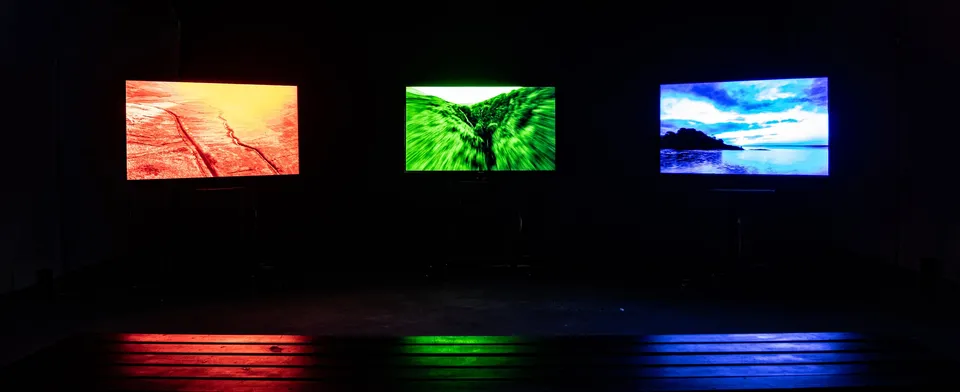 Three screens in a dark room glowing red, green and blue, their colours reflected on the floor